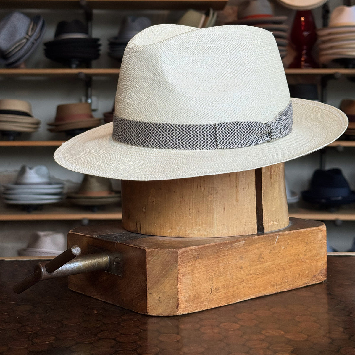 Italin Panama Fedora in light natural straw with a grey woven band, displayed on a wooden hat block in a shop with various hats in the background.