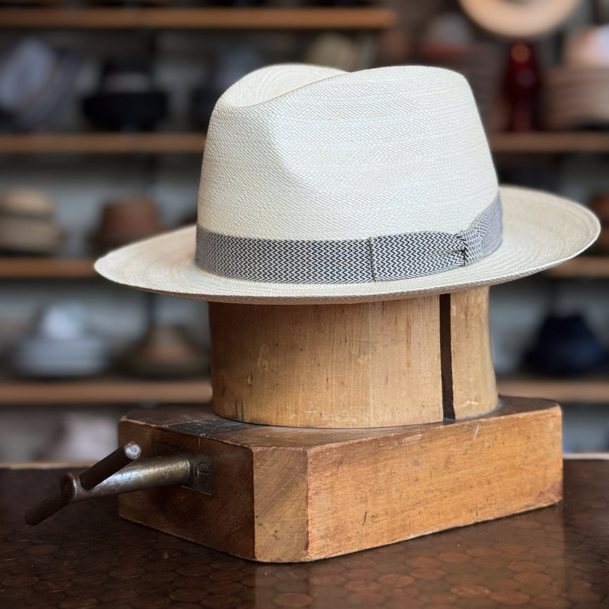 Italin Panama Fedora in light natural weave with a gray textured band, displayed on a vintage wooden hat block against a blurred workshop background.