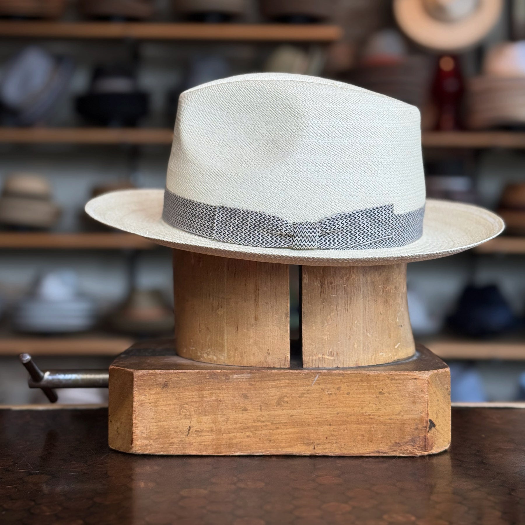 Italin Panama Fedora made of light woven material with a patterned gray band, displayed on a wooden hat block with shelves of hats in the background.