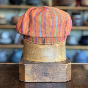 Juicy Hatteras newsboy cap featuring vibrant orange and multicolor stripes displayed on a vintage wooden hat block in a cozy hat shop setting.