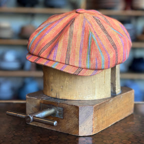 Juicy Hatteras newsboy cap in vibrant orange with multicolored stripes, displayed on a wooden hat block, showcasing its classic flat cap style and fabric texture.