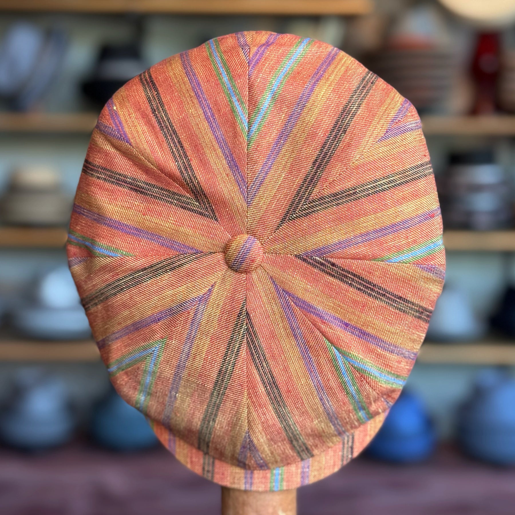 Juicy Hatteras flat cap with a vibrant orange base featuring multicolored striped patterns radiating from the center button, displayed on a wooden stand.