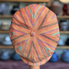 Juicy Hatteras flat cap with a vibrant orange base featuring multicolored striped patterns radiating from the center button, displayed on a wooden stand.