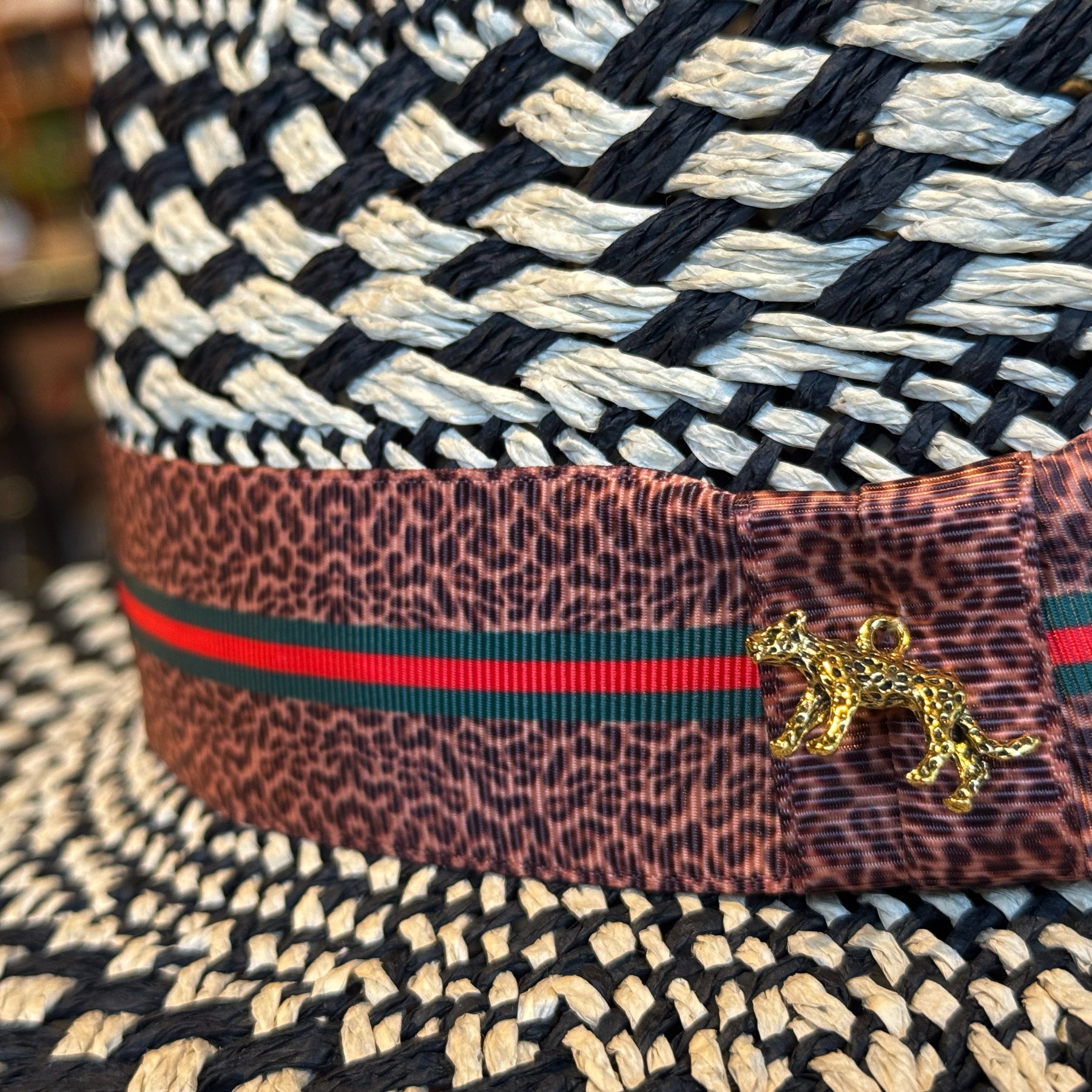Close-up of a woven black and ivory pattern hat with a brown leopard print band featuring green and red stripes and a small gold leopard charm, branded Kingston.