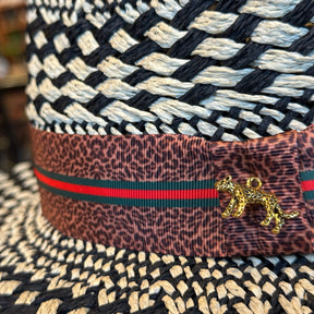 Close-up of a woven black and ivory pattern hat with a brown leopard print band featuring green and red stripes and a small gold leopard charm, branded Kingston.