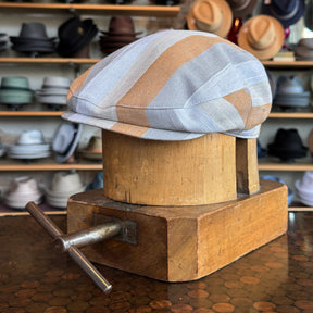 Kit Flat Cap with a striped pattern in soft beige, gray, and light blue tones, displayed on a vintage wooden hat block in a hat shop background.