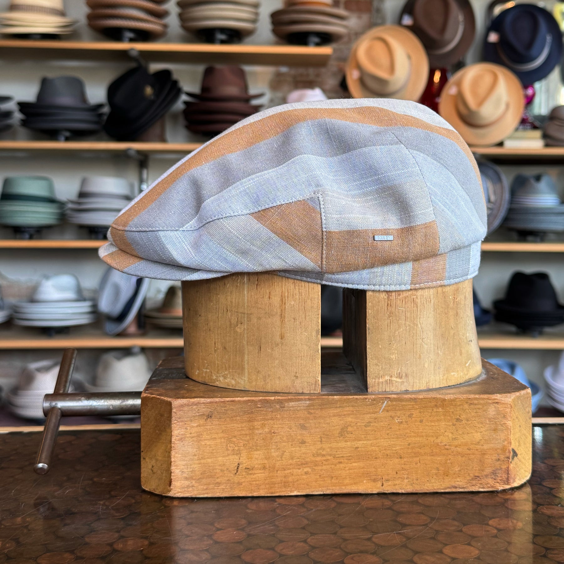 Kit Flat Cap in muted tones of grey, beige, and light blue displayed on a wooden hat block in a hat shop with various styles in the background.