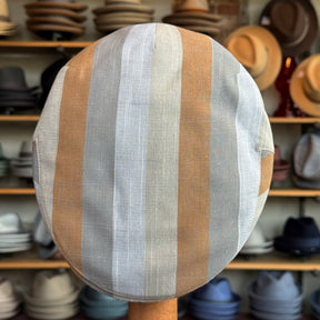 Top view of the Kit Flat Cap featuring wide vertical stripes in soft beige, light gray, and muted brown tones, displayed in a hat shop background with various hats on shelves.
