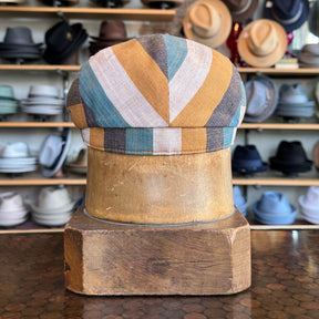 Kit Flat Cap in multicolor striped fabric displayed on a wooden hat block with shelves of various hats in the background.