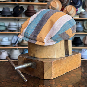 Kit Flat Cap with multicolored stripes in tan, blue, brown, and cream displayed on a wooden hat block in a hat shop featuring various hats in the background.