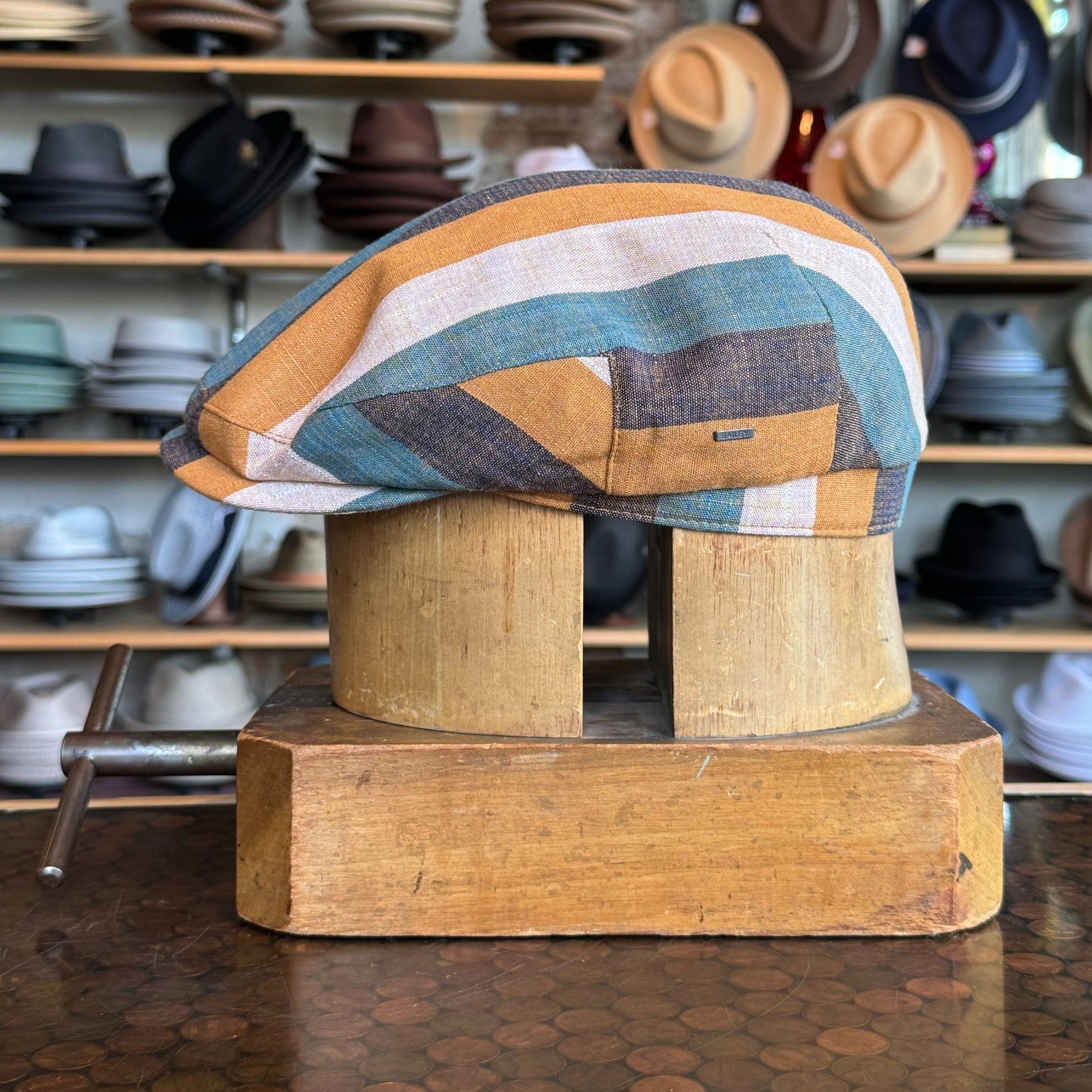 Kit Flat Cap featuring a multicolored striped pattern with shades of brown, blue, and cream, displayed on a wooden hat block in a hat shop background.