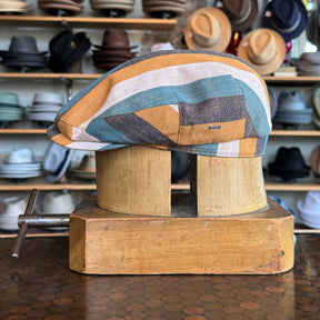 Kit Flat Cap featuring a multicolored striped pattern with shades of brown, blue, and cream, displayed on a wooden hat block in a hat shop background.