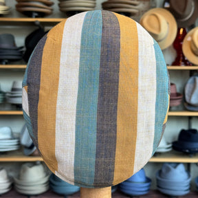 Kit Flat Cap with vertical stripes in muted shades of blue, yellow, brown, and cream, displayed in a hat store with various hats in the background.