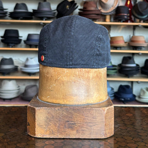 Leland Two-Panel Ivy Cap in dark fabric displayed on a wooden hat block with shelves of various styles of hats blurred in the background.