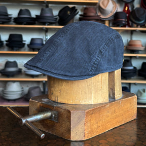 Leland Two-Panel Ivy Cap in dark fabric displayed on a wooden hat block with a background of various hats on shelves in a hat shop.