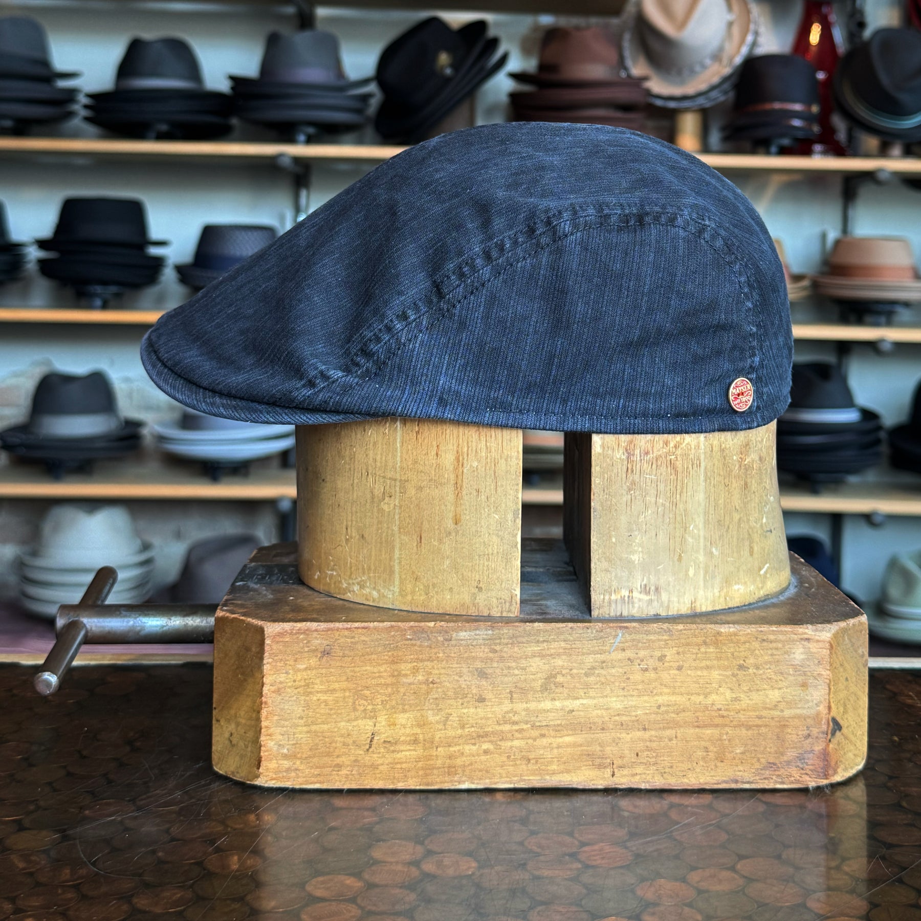 Leland Two-Panel Ivy Cap in dark fabric displayed on a wooden hat block with a variety of hats in the blurred background in a stylish hat shop setting.