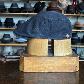 Leland Two-Panel Ivy Cap in dark fabric displayed on a wooden hat block with a variety of hats in the blurred background in a stylish hat shop setting.