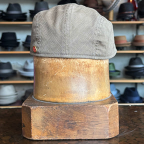 Leland Two-Panel Ivy Cap in khaki fabric displayed on a wooden hat block with various hats blurred in the background, showcasing a classic flat cap style.