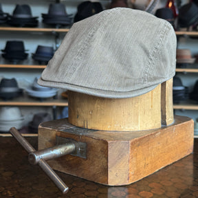 Light khaki fabric Leland Two-Panel Ivy Cap displayed on a vintage wooden hat block with shelves of various hats blurred in the background.