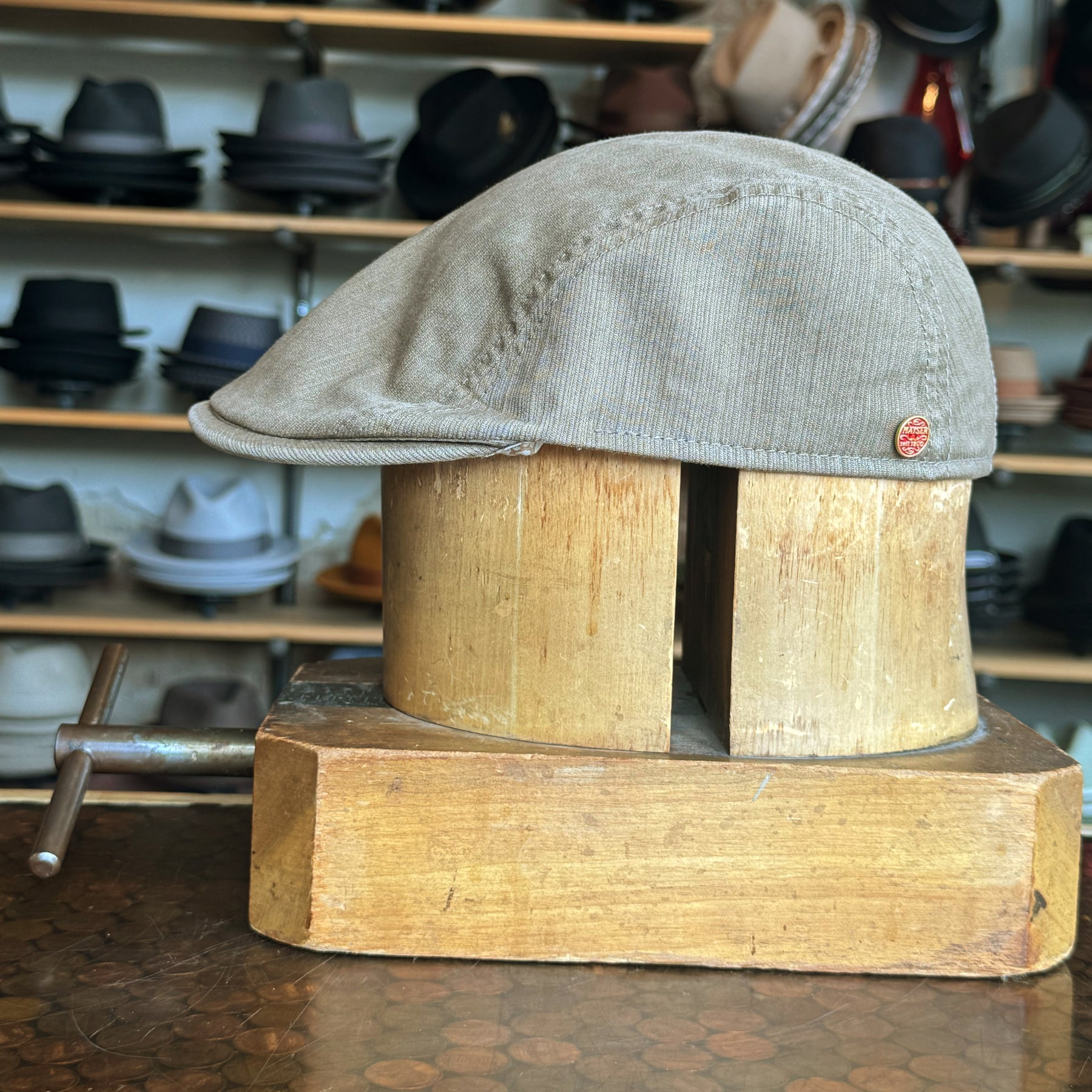 Side view of a khaki Leland Two-Panel Ivy Cap displayed on a wooden hat block with shelves of various hats blurred in the background.
