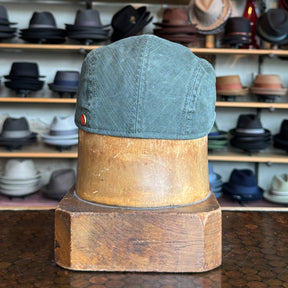 Back view of the Leland Two-Panel Ivy Cap in muted green fabric displayed on a wooden hat block with shelves of various hats in the background.