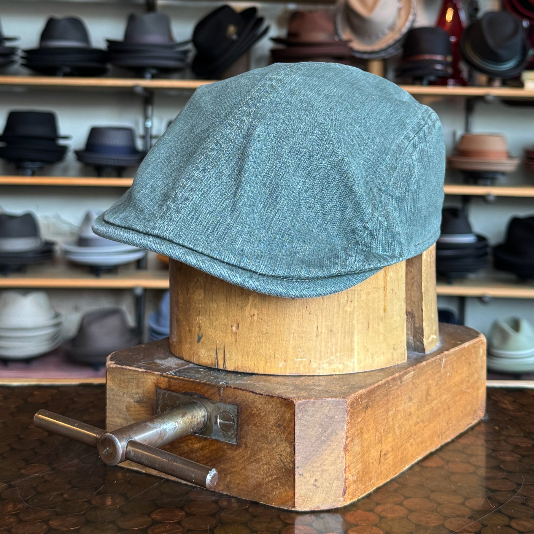 Leland Two-Panel Ivy Cap in muted green fabric displayed on a wooden hat block with various hats in the background, showcasing a classic flat cap style.