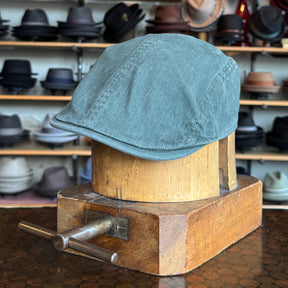 Leland Two-Panel Ivy Cap in muted green fabric displayed on a wooden hat block with various hats in the background, showcasing a classic flat cap style.
