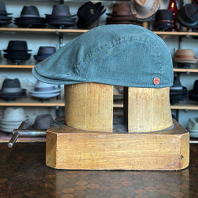 Leland Two-Panel Ivy Cap in soft textured fabric displayed on a wooden hat block with shelves of various brim hats in the background.