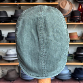 Leland Two-Panel Ivy Cap in olive green textured fabric displayed on a stand with shelves of various hats in the background.