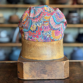 Liberty Deco Flat Cap displayed on a wooden hat block, featuring a vibrant multicolor floral and geometric pattern with a soft fabric construction.