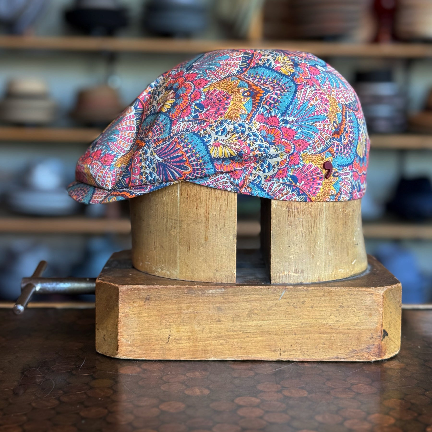 Colorful patterned Liberty Deco Flat Cap displayed on a wooden hat block, featuring an intricate floral and abstract design with vibrant reds, blues, and yellows.