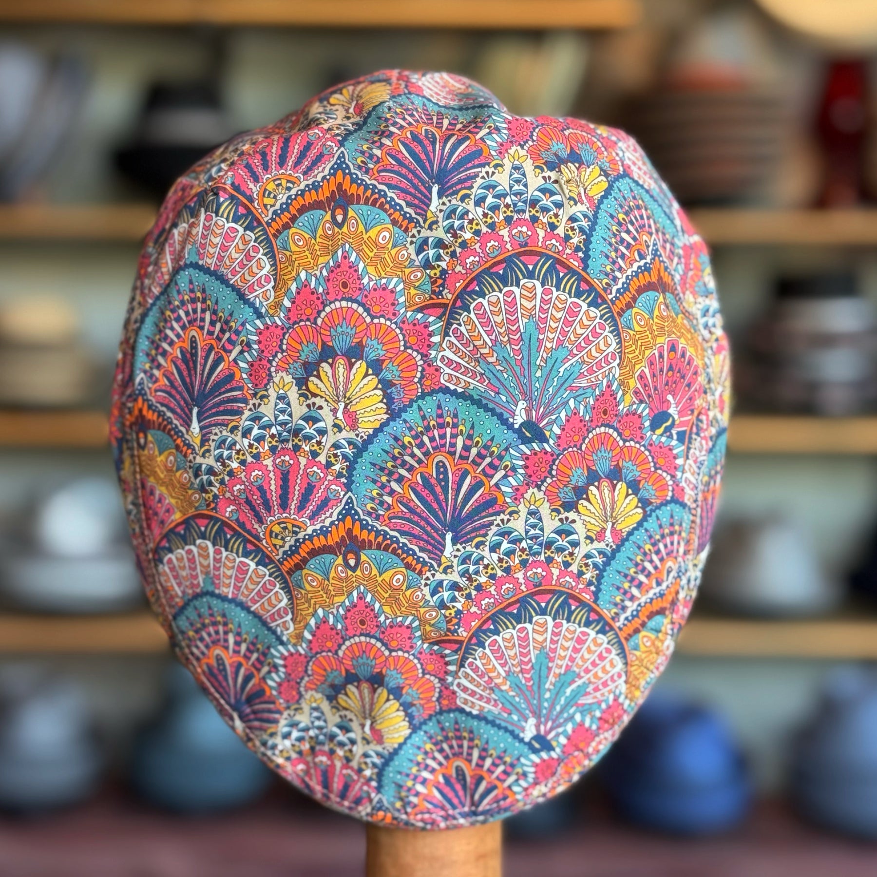Liberty Deco Flat Cap featuring an intricate multicolored fan and floral pattern with vibrant pink, blue, yellow, and orange hues, displayed against a blurred background of shelves and pottery.