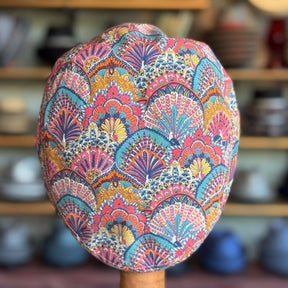 Liberty Deco Flat Cap featuring an intricate multicolored fan and floral pattern with vibrant pink, blue, yellow, and orange hues, displayed against a blurred background of shelves and pottery.
