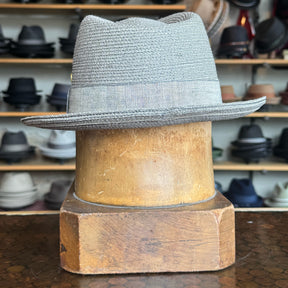 Linen Maleo Fedora in light gray with a woven texture and matching fabric band, displayed on a wooden hat block with shelves of various hats in the background.