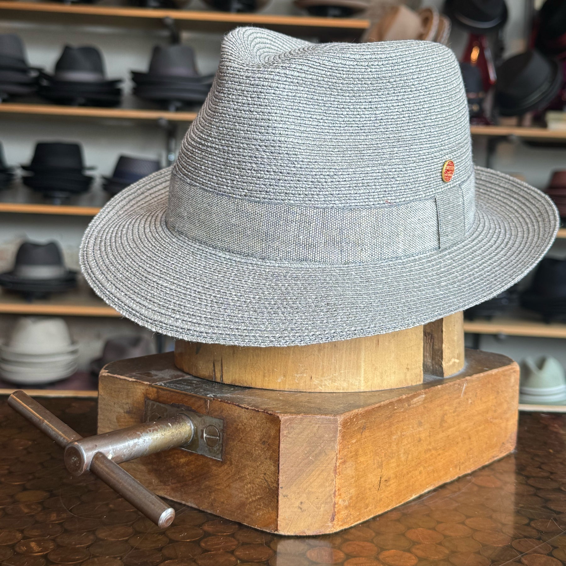 Elegant Linen Maleo Fedora in light gray woven material displayed on a wooden hat block with various hats in the background, showcasing classic brim style.