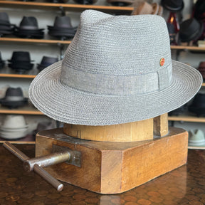 Elegant Linen Maleo Fedora in light gray woven material displayed on a wooden hat block with various hats in the background, showcasing classic brim style.