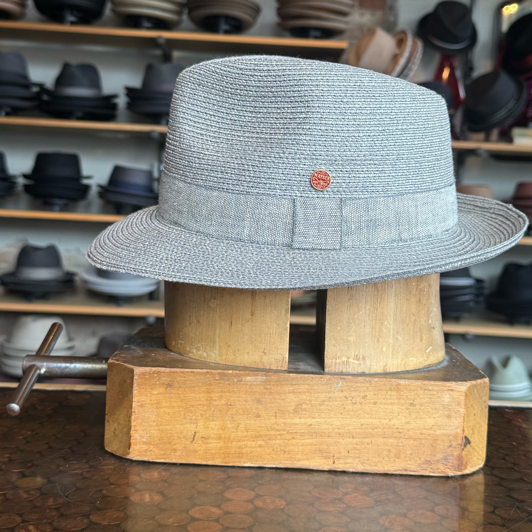 Linen Maleo Fedora in light gray linen fabric with a classic ribbon band and small red emblem, displayed on a wooden hat block in a store with shelves of various hats in the background.