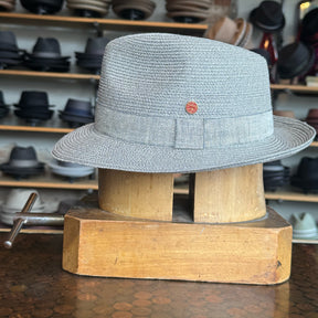 Linen Maleo Fedora in light gray linen fabric with a classic ribbon band and small red emblem, displayed on a wooden hat block in a store with shelves of various hats in the background.