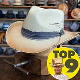 Loredo straw hat with a wide brim and a dark fabric band featuring a decorative metal emblem, displayed on a wooden hat block in a hat shop.