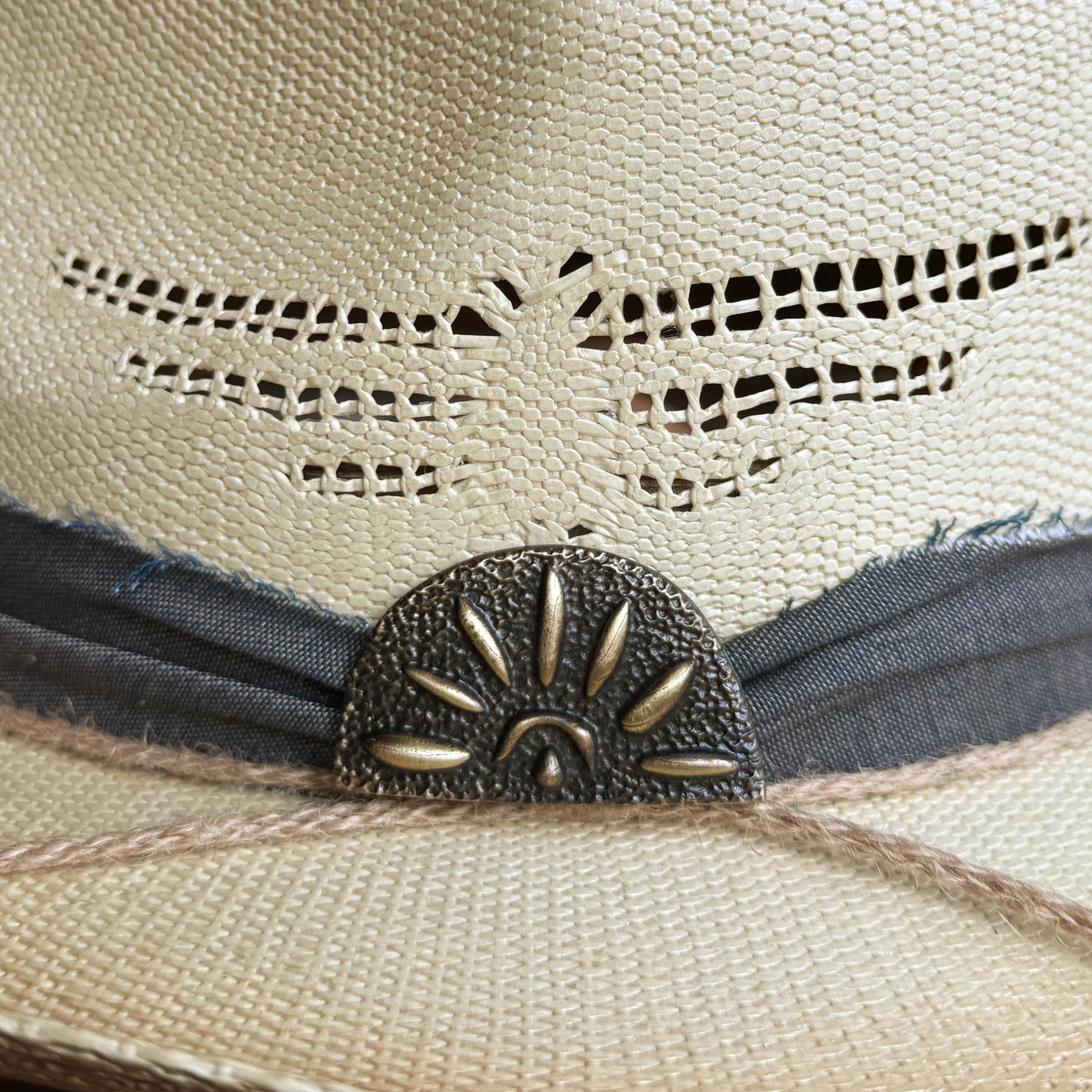 Close-up view of the woven detail and decorative metal accent on the woven hat named Loredo, featuring a distressed fabric band and natural fibers.