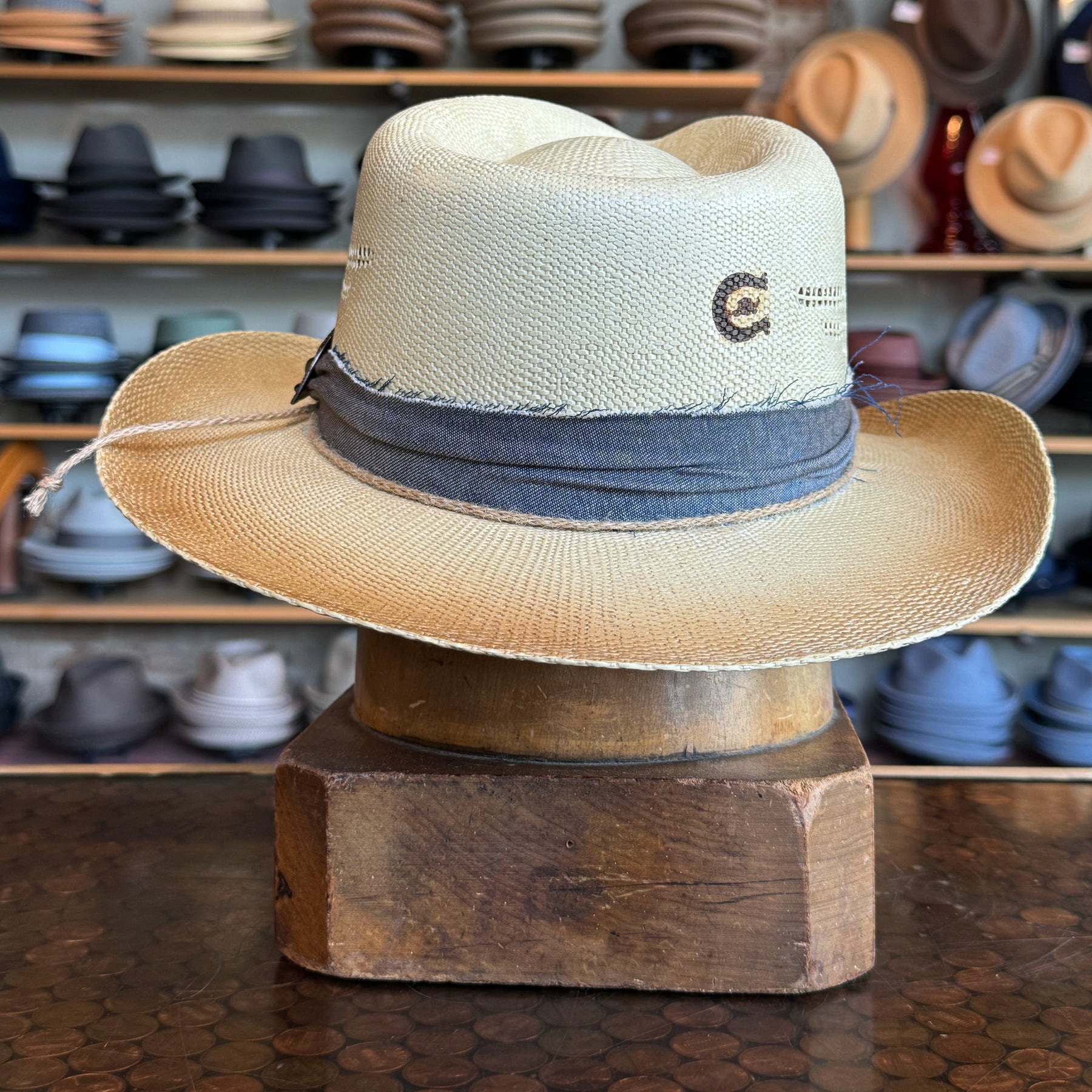 Loredo straw hat with a wide brim and gray fabric band detail, displayed on a wooden stand with various hats blurred in the background.