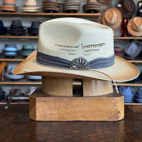 Loredo hat in light tan straw with dark brown band and decorative concho, displayed on wooden hat block with various hats blurred in background.