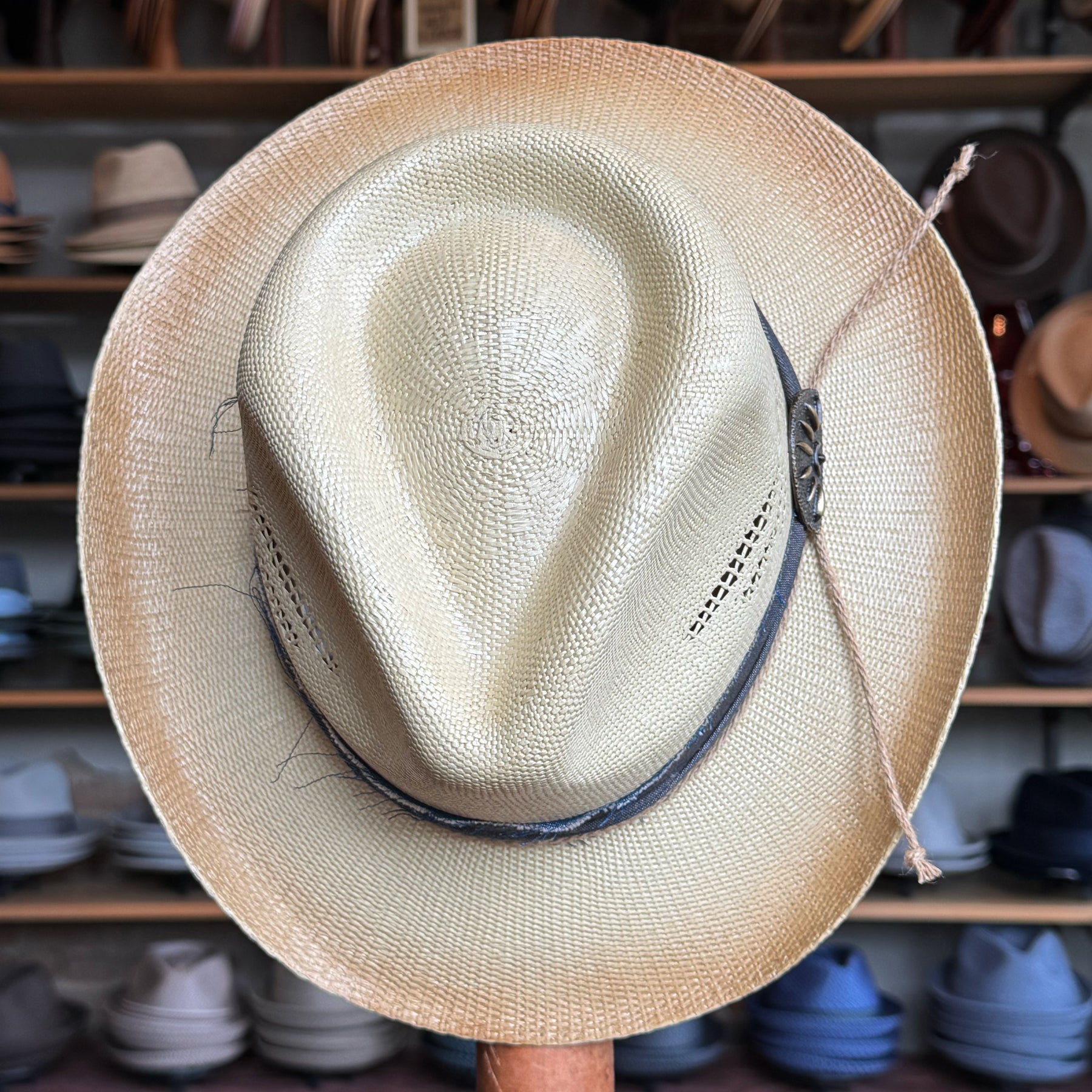 Loredo straw brim hat featuring a wide natural-colored brim and a textured crown with a dark leather band and decorative side tie, displayed in a hat shop background.