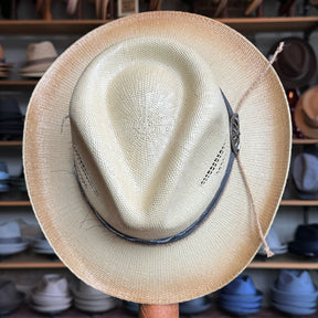 Loredo straw brim hat featuring a wide natural-colored brim and a textured crown with a dark leather band and decorative side tie, displayed in a hat shop background.