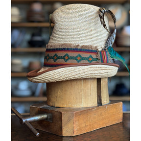 Luna Straw Top Hat featuring woven straw material with a decorative multicolored band and patterned feathers, displayed on a vintage wooden hat block.