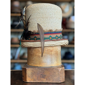 Luna Straw Top Hat featuring a woven straw design with a colorful patterned band and decorative feathers, displayed on a wooden hat block in a vintage setting.