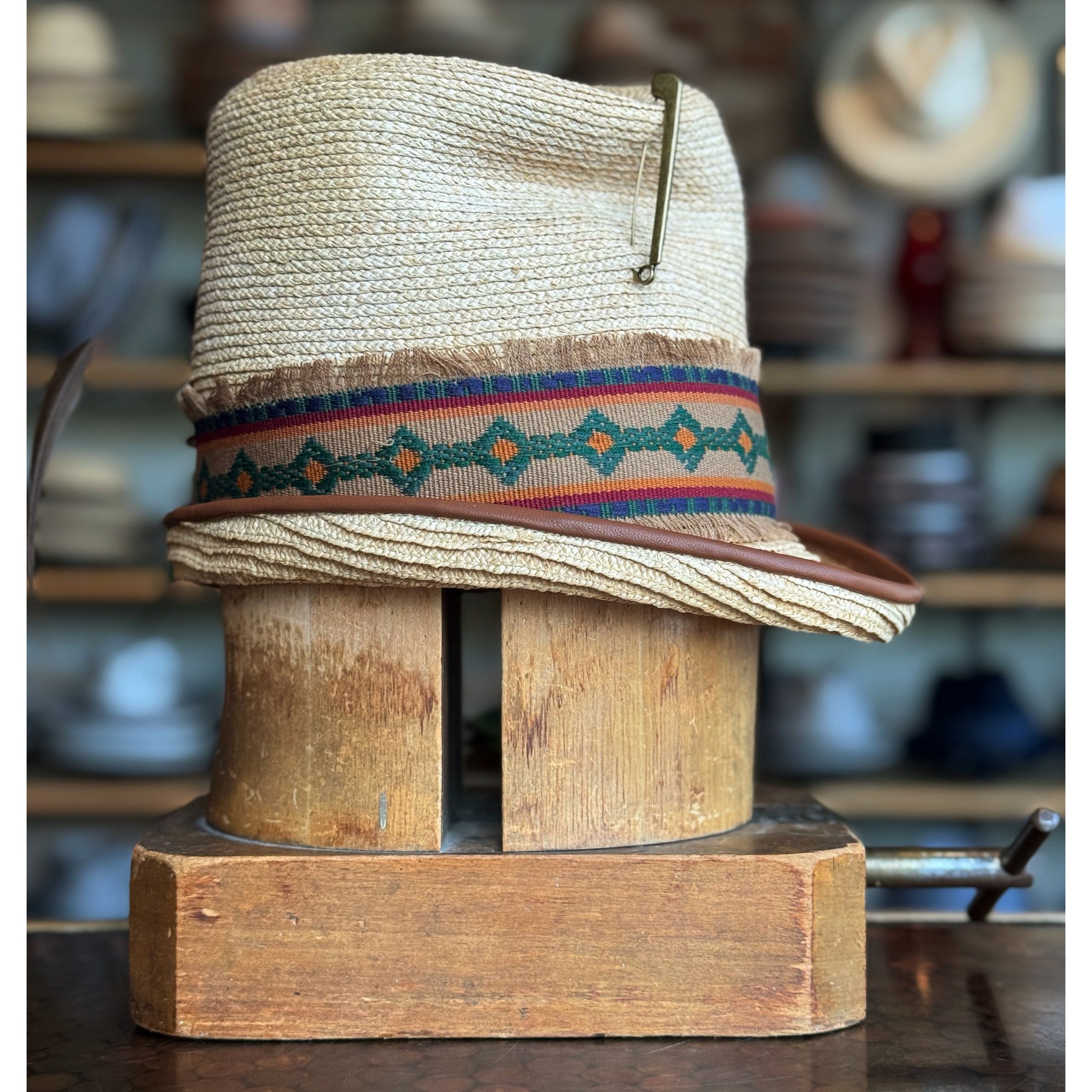 Luna Straw Top Hat featuring woven straw material with a colorful patterned fabric band and a decorative pin, displayed on a wooden hat block in a vintage setting.
