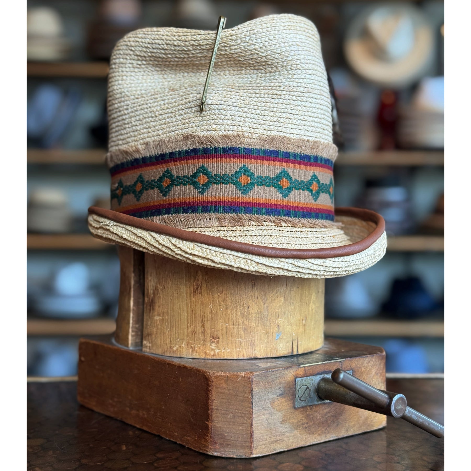 Luna Straw Top Hat featuring a woven straw design with a patterned fabric band in earthy tones, displayed on a wooden hat block in a vintage-style setting.