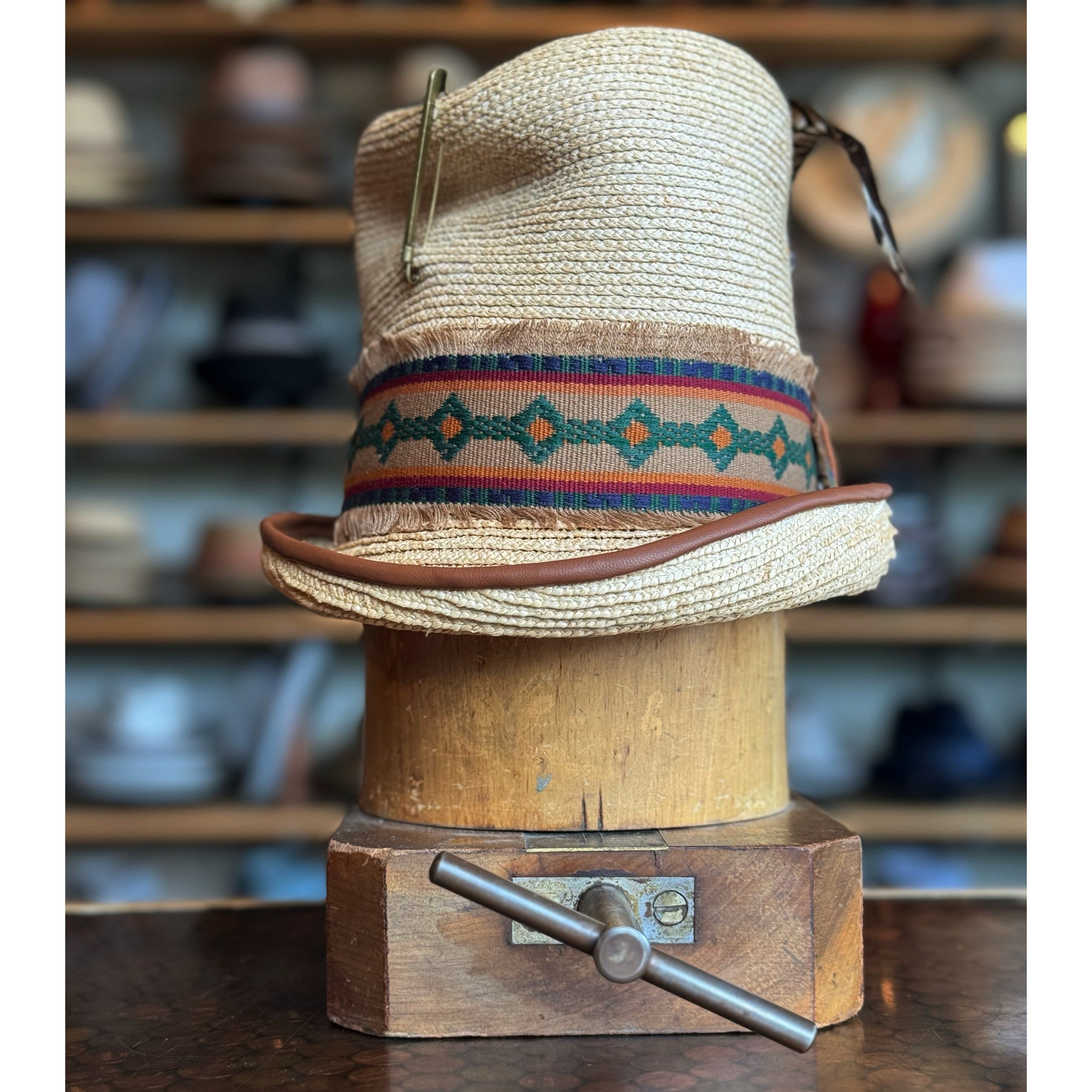 Luna Straw Top Hat made from woven straw with a colorful patterned band and a slightly curved brim, displayed on a wooden hat block in a rustic setting.