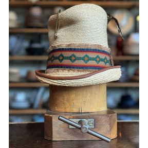 Luna Straw Top Hat made from woven straw with a colorful patterned band and a slightly curved brim, displayed on a wooden hat block in a rustic setting.